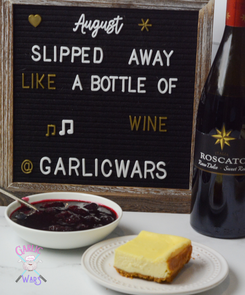 cheesecake bar on small white plate with red wine berry sauce in small white bowl, in front of letterboard that reads lyrics from a Taylor Swift song "august slipped away like a bottle of wine"