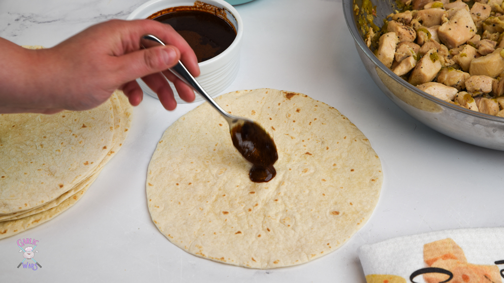 spoonful of enchilada sauce being poured on tortilla