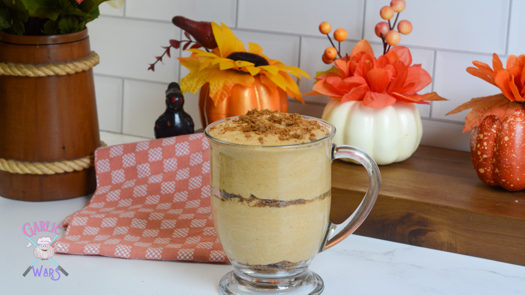 large glass cup filled with pumpkin mousse, layered with crushed ginger snaps, with small pumpkins and fall themed flowers in the background