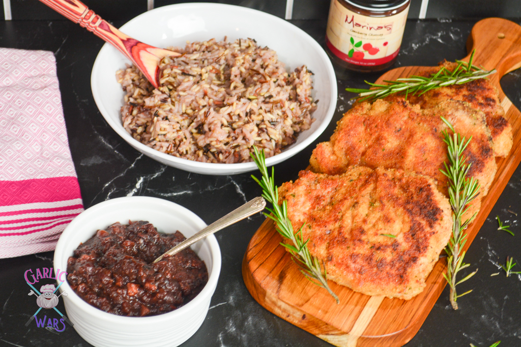 family style dinner - pork chops on a cutting board with rosemary, a bowl of wild rice, and a bowl of cranberry chutney