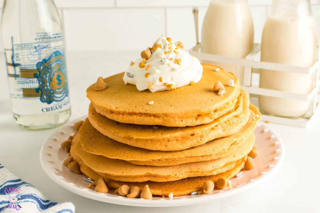 big stack of pancakes on a white plate topped with whipped cream and sprinkles